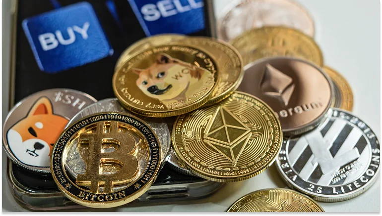 A variety of physical cryptocurrency coins including Bitcoin, Ethereum, Litecoin, and Dogecoin placed on a smartphone screen displaying buy and sell buttons.