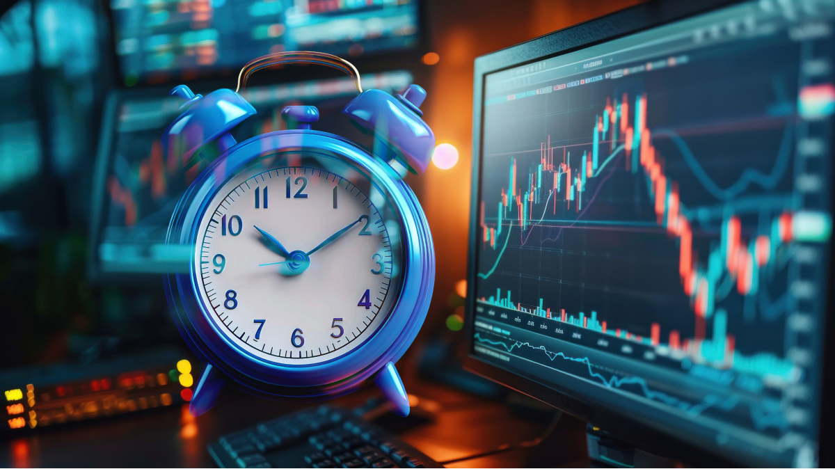 Photo of a blue vintage-style alarm clock placed on a trading desk beside multiple computer monitors showing candlestick price charts and technical indicators.
