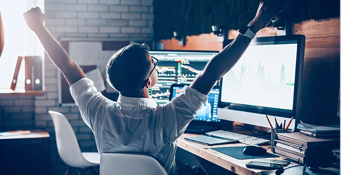 Excited trader celebrating success at desk with multiple monitors showing stock charts