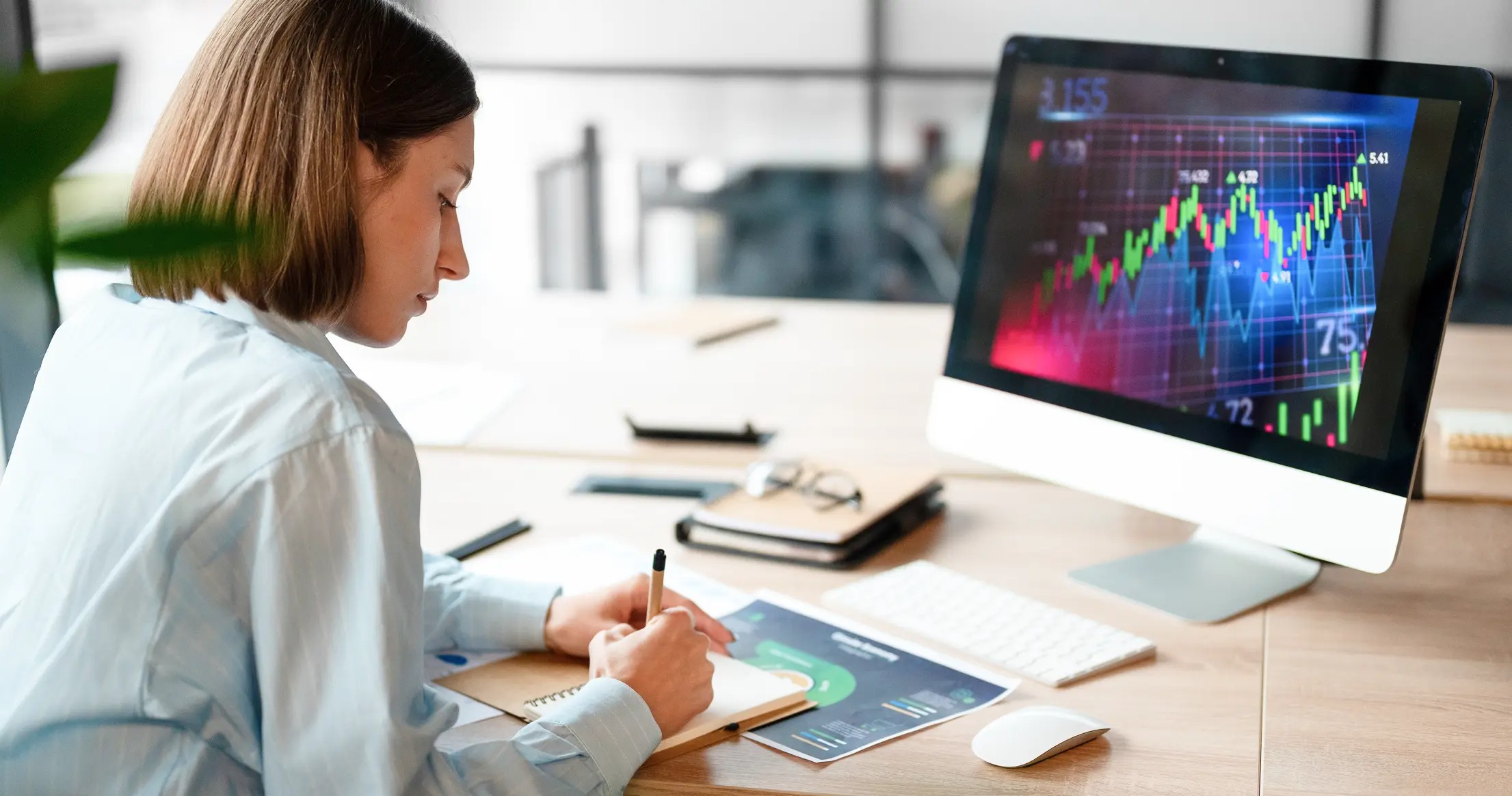 Woman taking notes at a desk while a desktop monitor displays a candlestick chart, representing market analysis and trading study